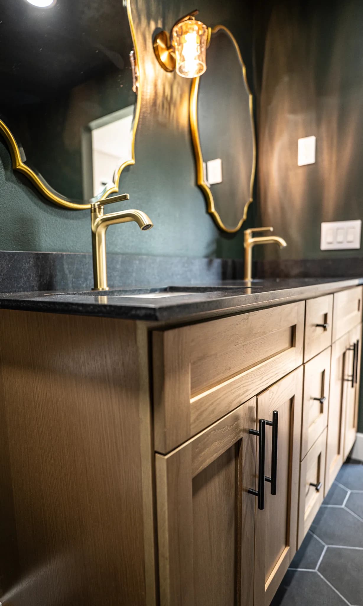Green bathroom with oak vanity cabinets, a black counter, and gold faucets and mirrors