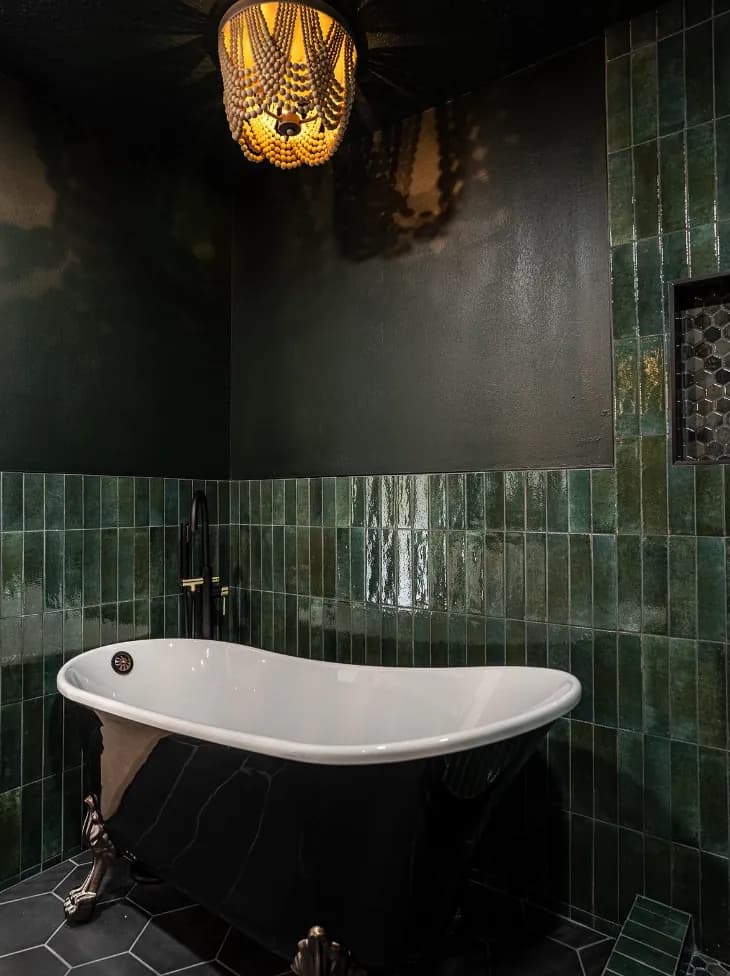 Black clawfoot tub, green tile wainscot, dark walls, gold chandelier