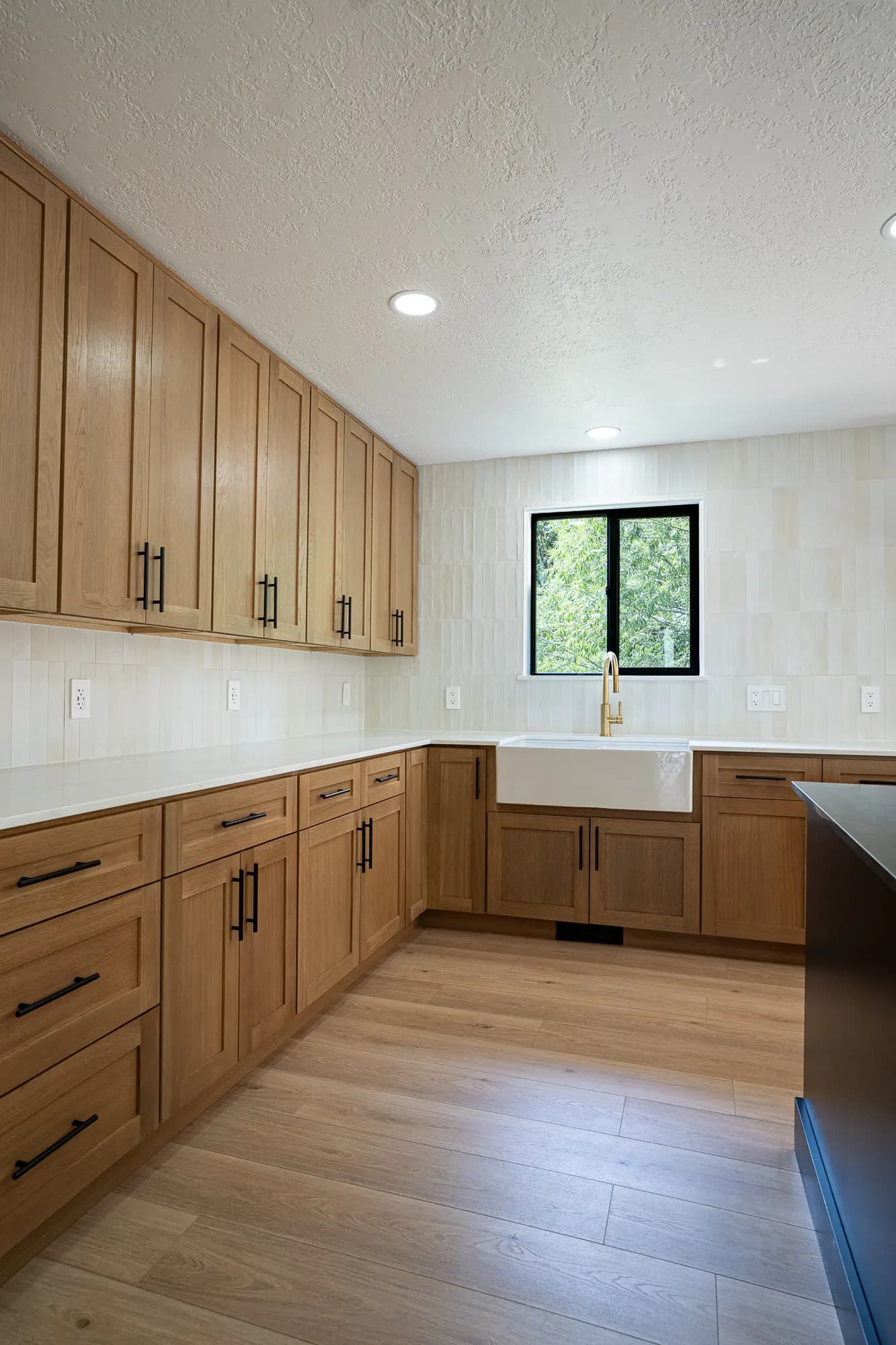 White oak corner with a farmhouse sink, brass faucet, black pulls, and cream stacked tile